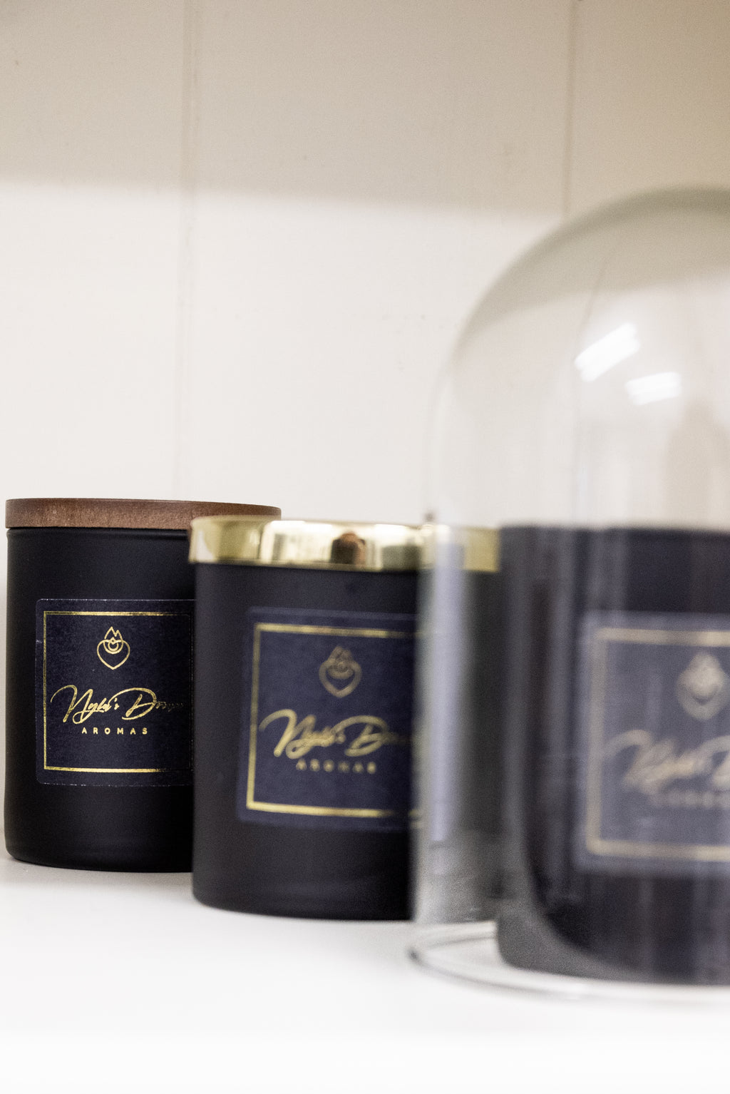 Three black candles with gold labels on a white surface, one under a glass dome.